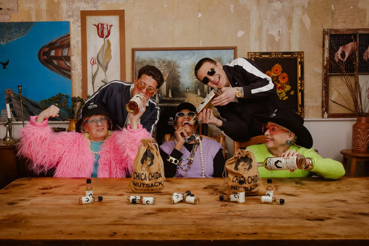 Three grandmothers and two men in tracksuits celebrate with Chica~Chida Nutsack burlap sacks and shooter bottles on a wooden table