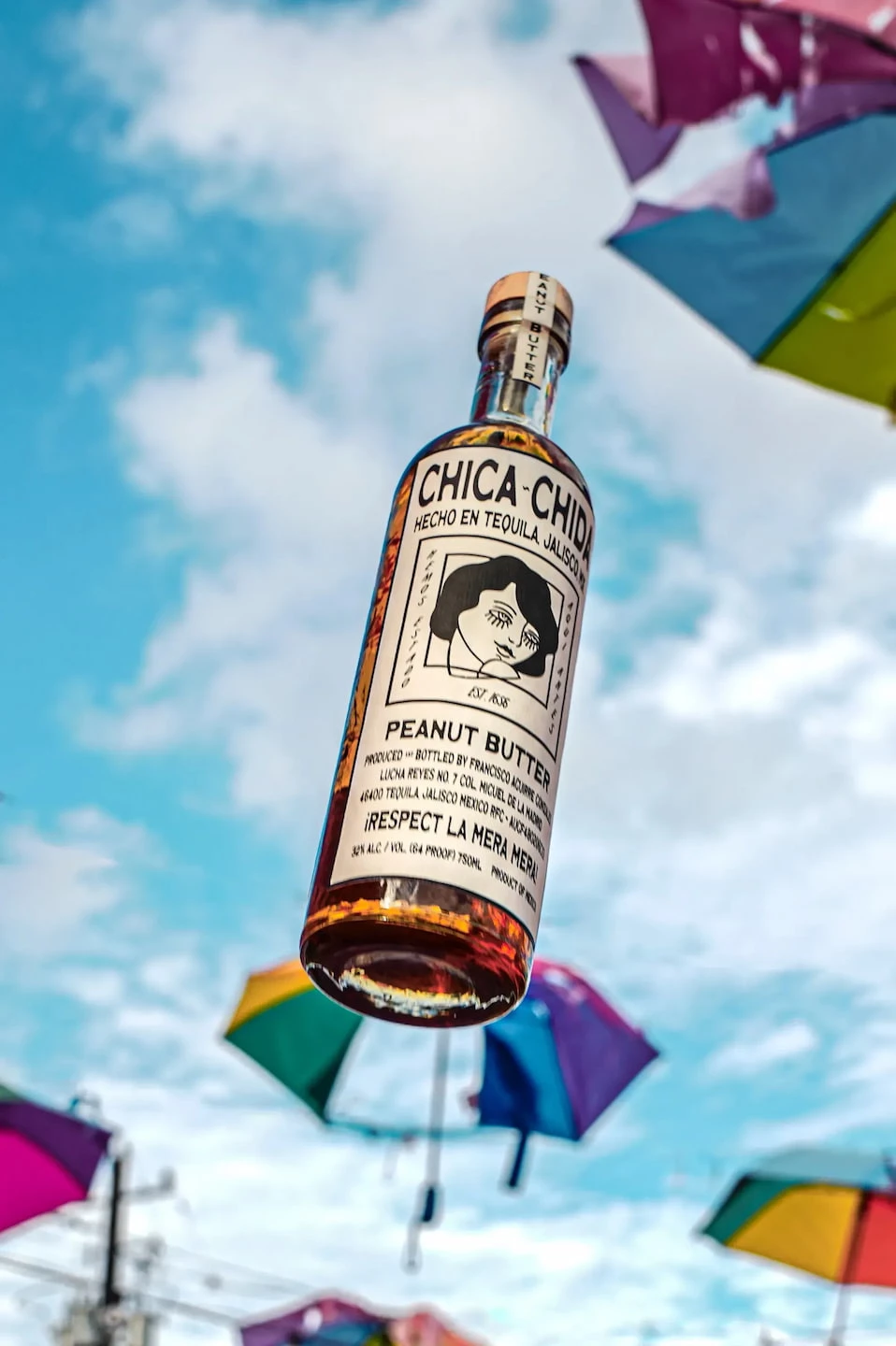 Chica~Chida peanut butter agave spirit bottle floating against a blue sky surrounded by colorful umbrellas