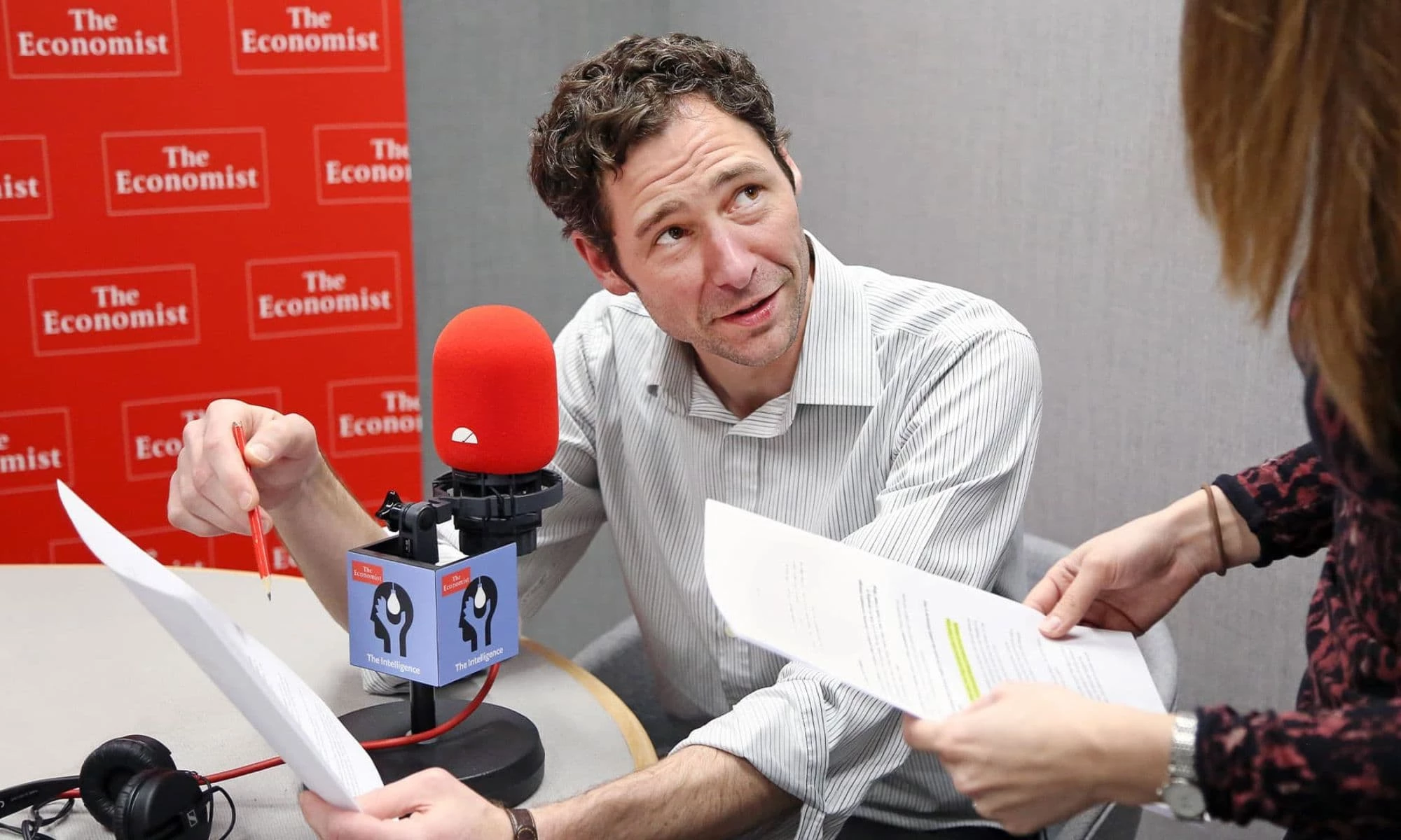 A journalist reviews notes during a recording session at The Economist's podcast studio, with The Intelligence branded microphone and red Economist backdrop visible