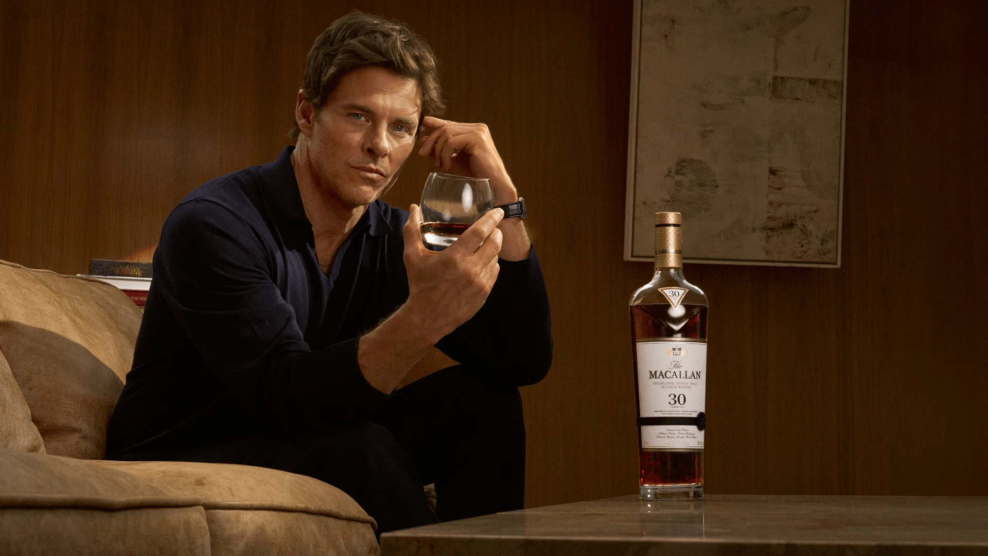 The Macallan Highlights Sherry Oak 30 in ‘Drink of a Generation’ James Marsden holds a whisky glass beside a bottle of The Macallan Sherry Oak 30 in a warm wood-paneled room.