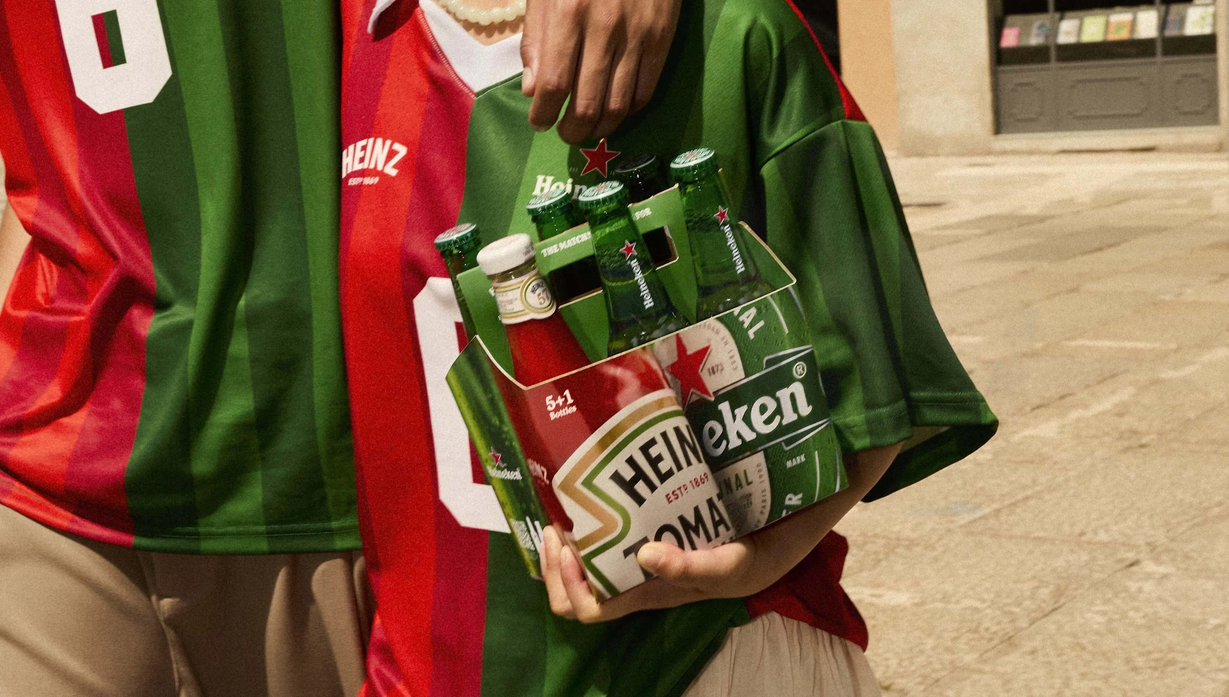 People in red-and-green soccer jerseys hold the Heinz x Heineken six-pack, featuring five Heineken beers and one Heinz ketchup bottle.