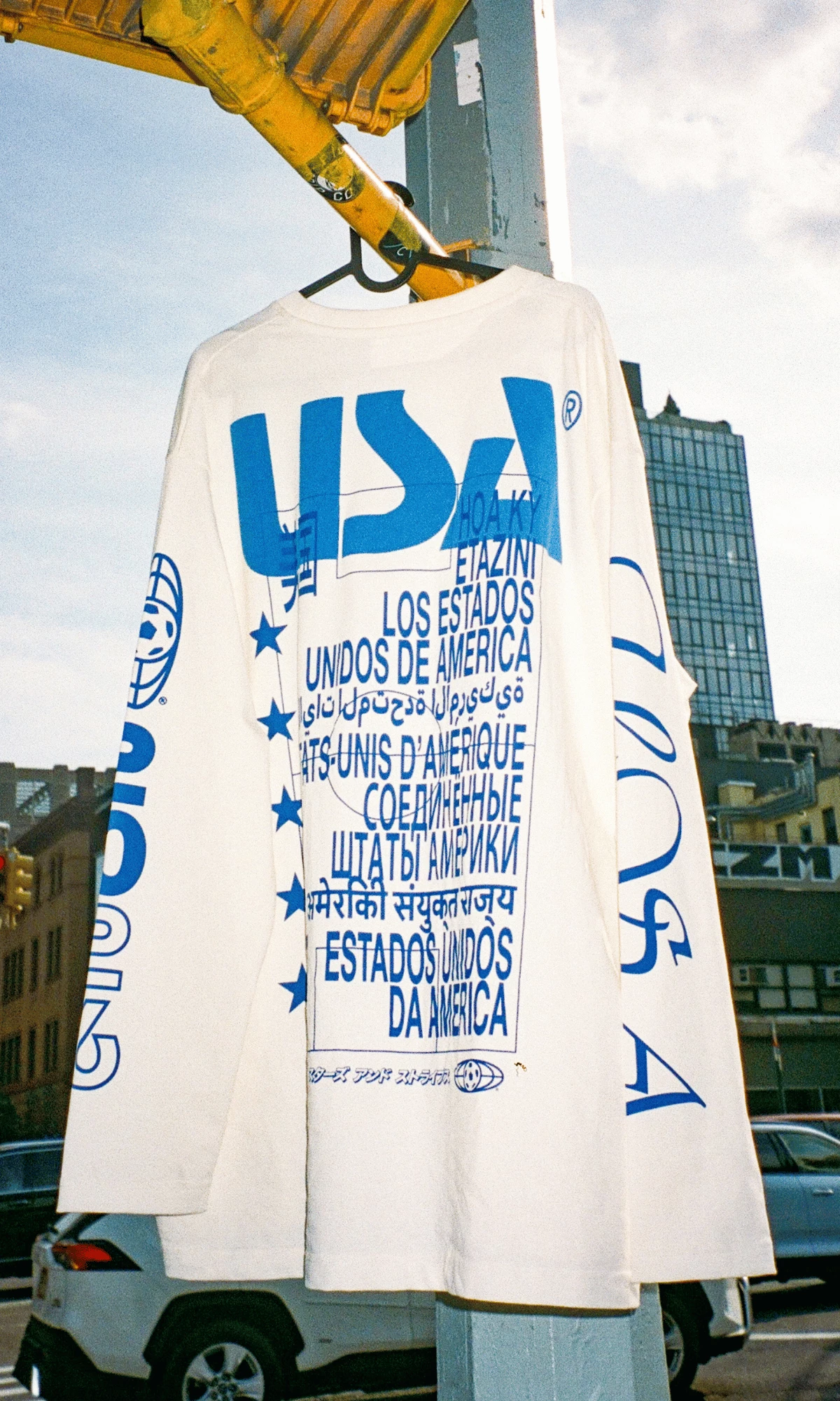 WIGZ’s white Immigrant Jersey hangs outdoors, showing “USA” in overlapping blue multilingual typography. WIGZ’s white Immigrant Jersey hangs outdoors, showing “USA” in overlapping blue multilingual typography.