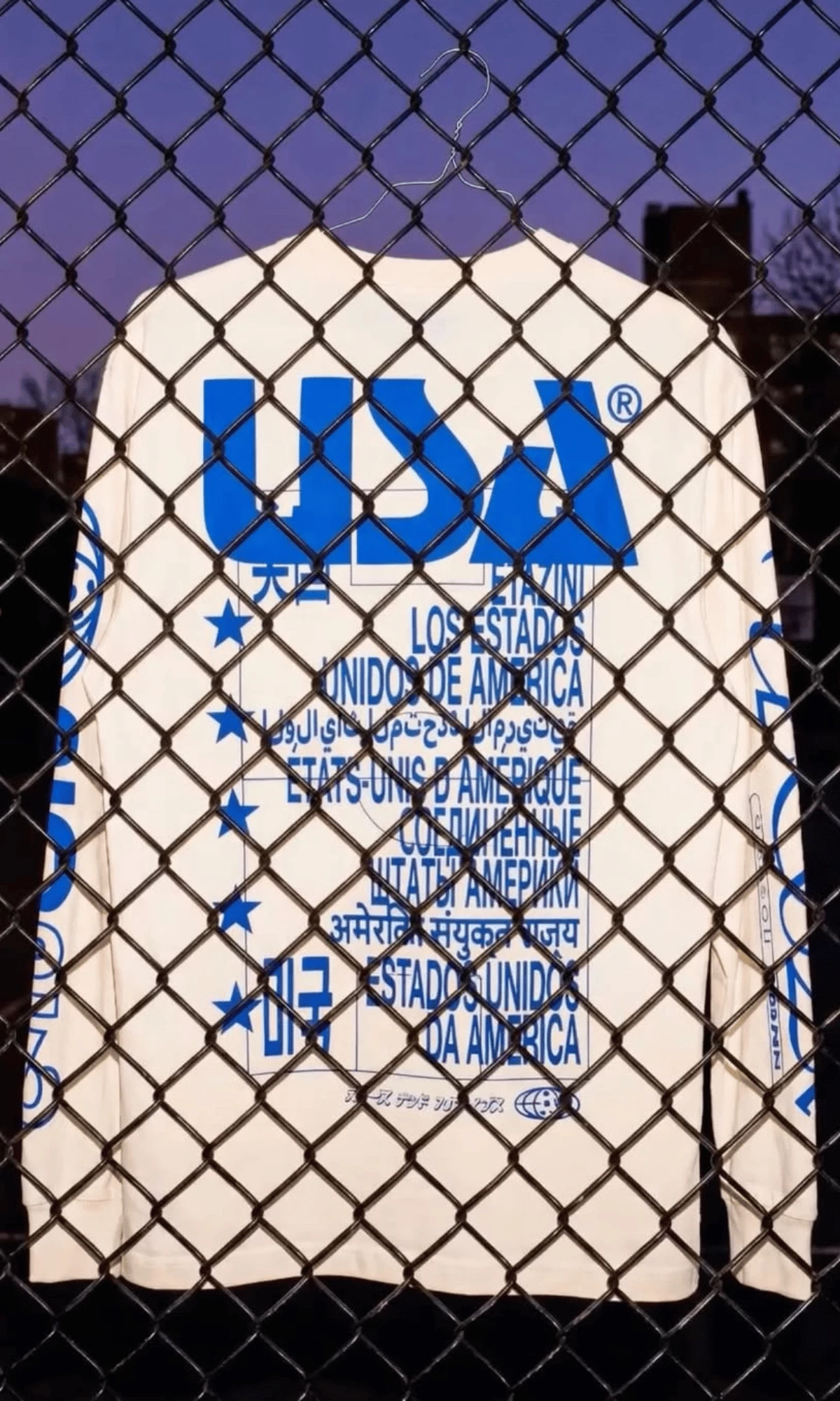 WIGZ Frames the Immigrant Jersey as a World Cup Statement WIGZ’s white Immigrant Jersey hangs behind a chain-link fence, with blue multilingual “USA” typography across the back.