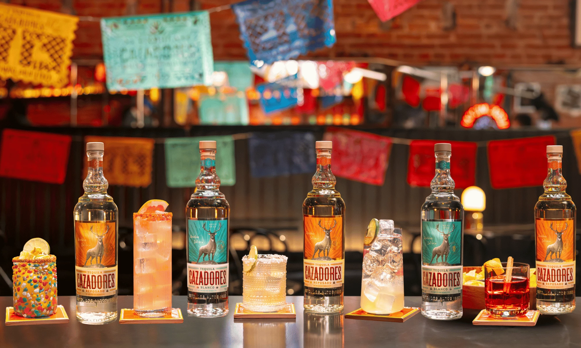 CAZADORES Pairs Fan Types With Matchday Cocktails Tequila CAZADORES bottles stand beside a lineup of colorful cocktails in a bar decorated with papel picado banners.