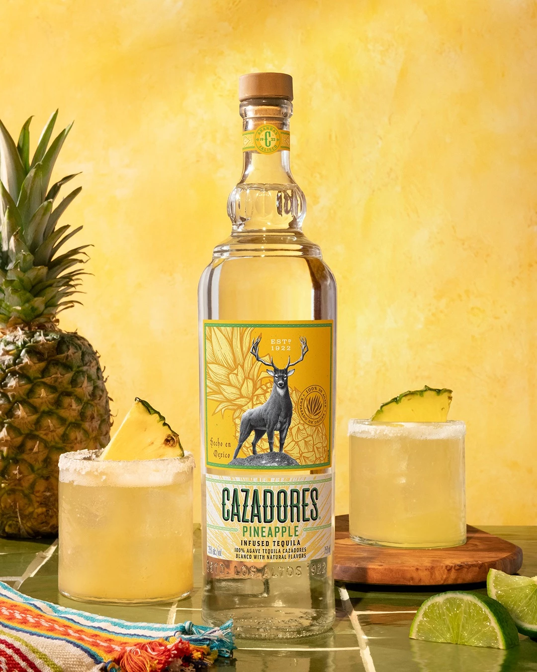 A bottle of CAZADORES Pineapple Infused Tequila stands between two yellow cocktails garnished with pineapple, alongside fresh pineapple and lime wedges.
