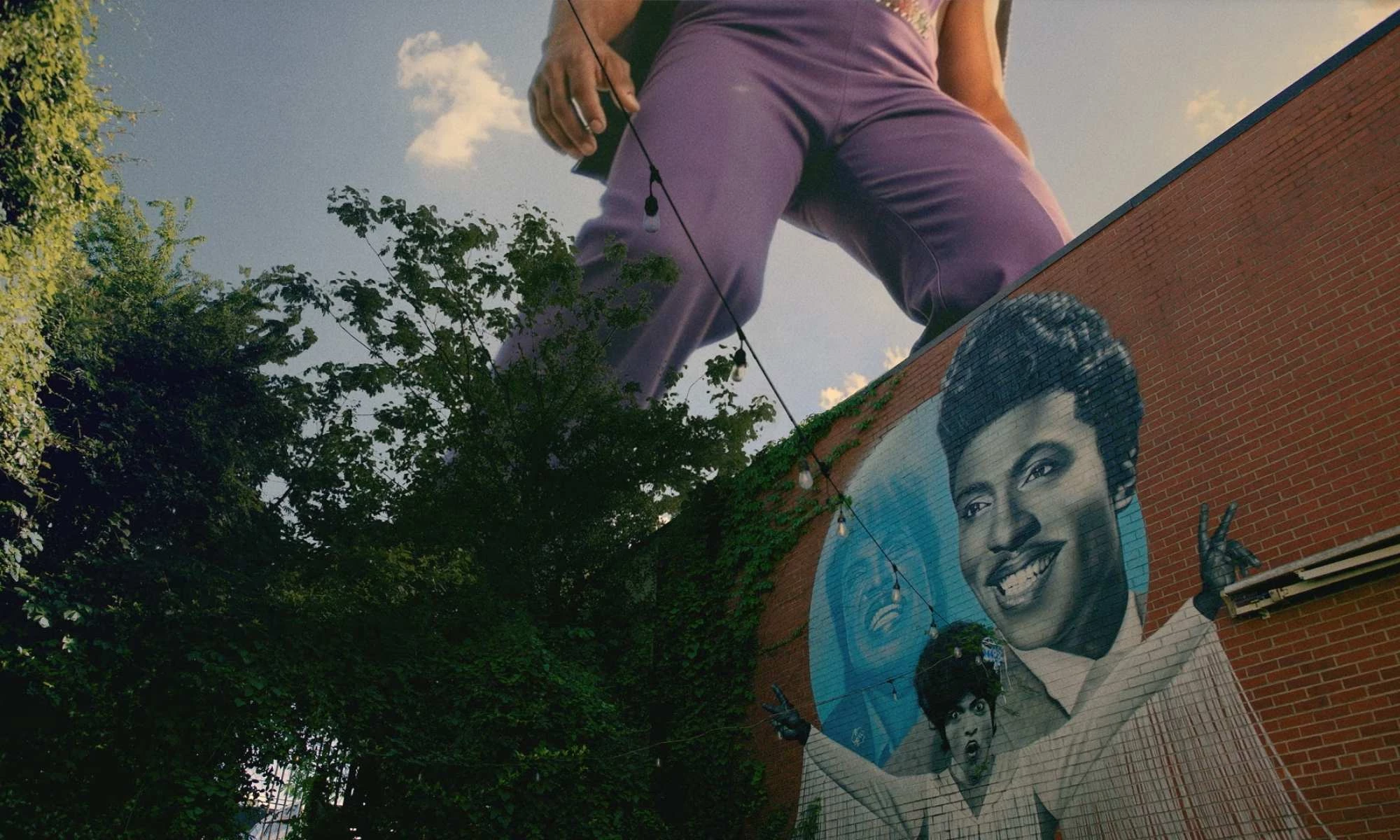 A larger-than-life figure in a purple outfit standing behind a Little Richard mural.