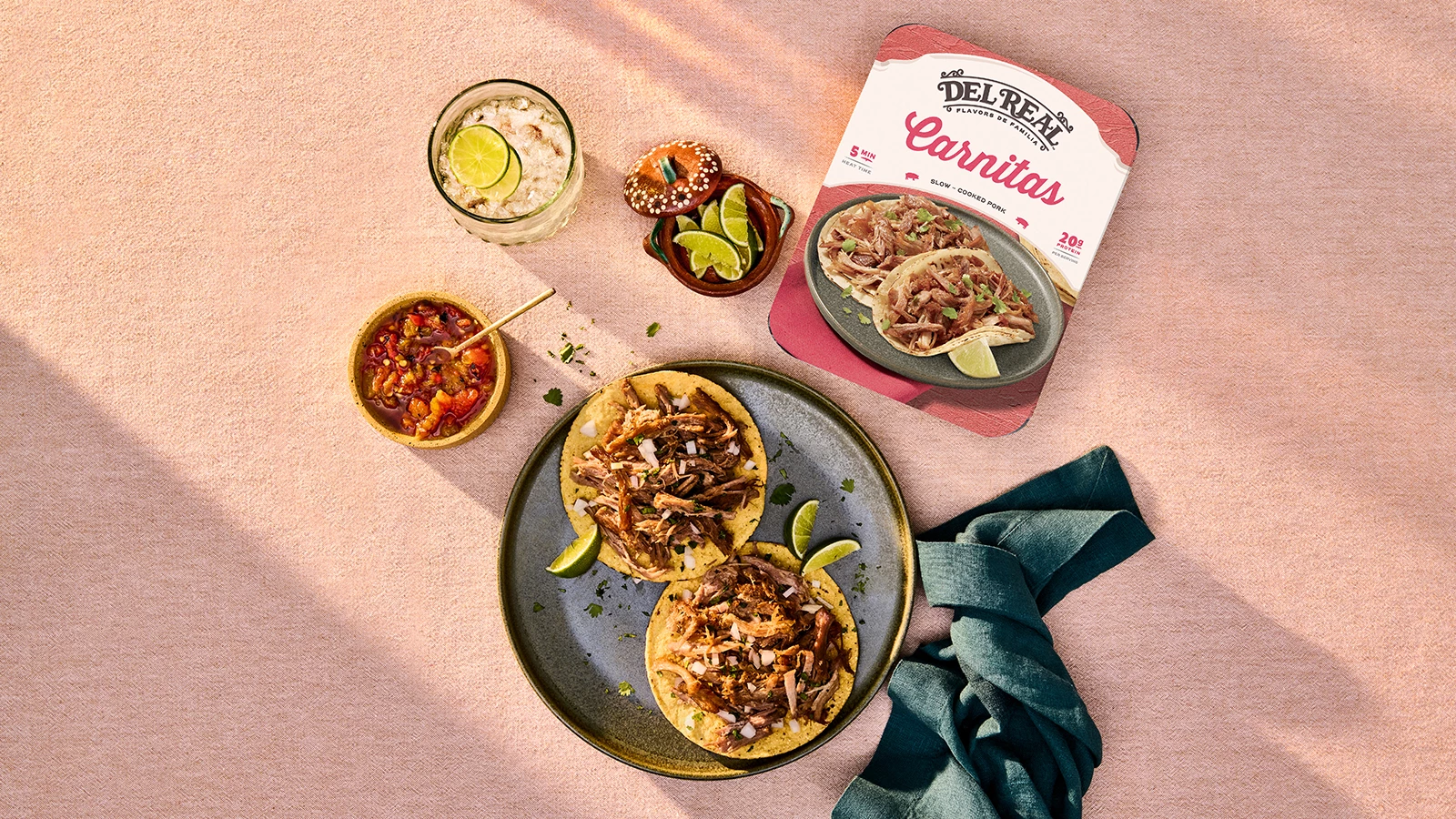 Del Real Foods carnitas tacos served with lime wedges, salsa, and a drink on a pink surface, alongside product packaging showing "Slow-Cooked Pork" ready in 5 minutes.