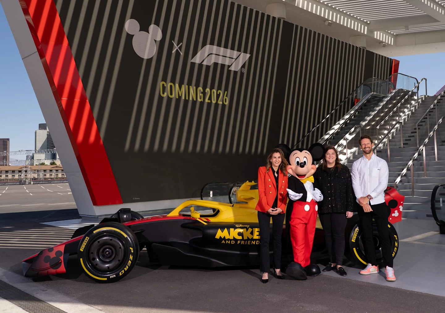 Tasia Filippatos, Mickey Mouse, Emily Prazer, and Romain Grosjean pose in front of a Disney x F1 race car at the Grand Prix Plaza in Las Vegas.