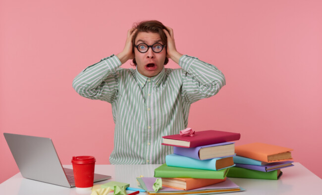 Person wearing round glasses holds their head in surprise at a desk with a laptop, coffee cup, and a colorful stack of books against a pink background.